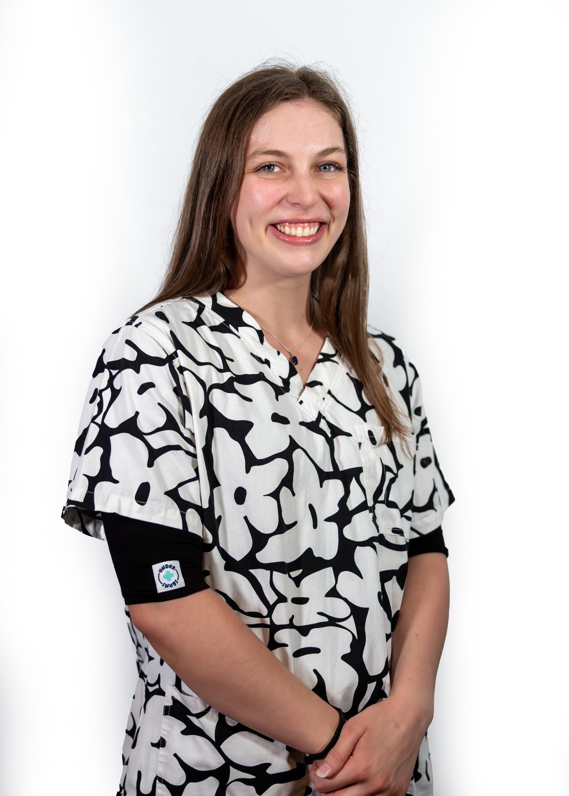 Smiling young nurse with long brown hair wearing black Under Snugs under her black & white floral scrubs