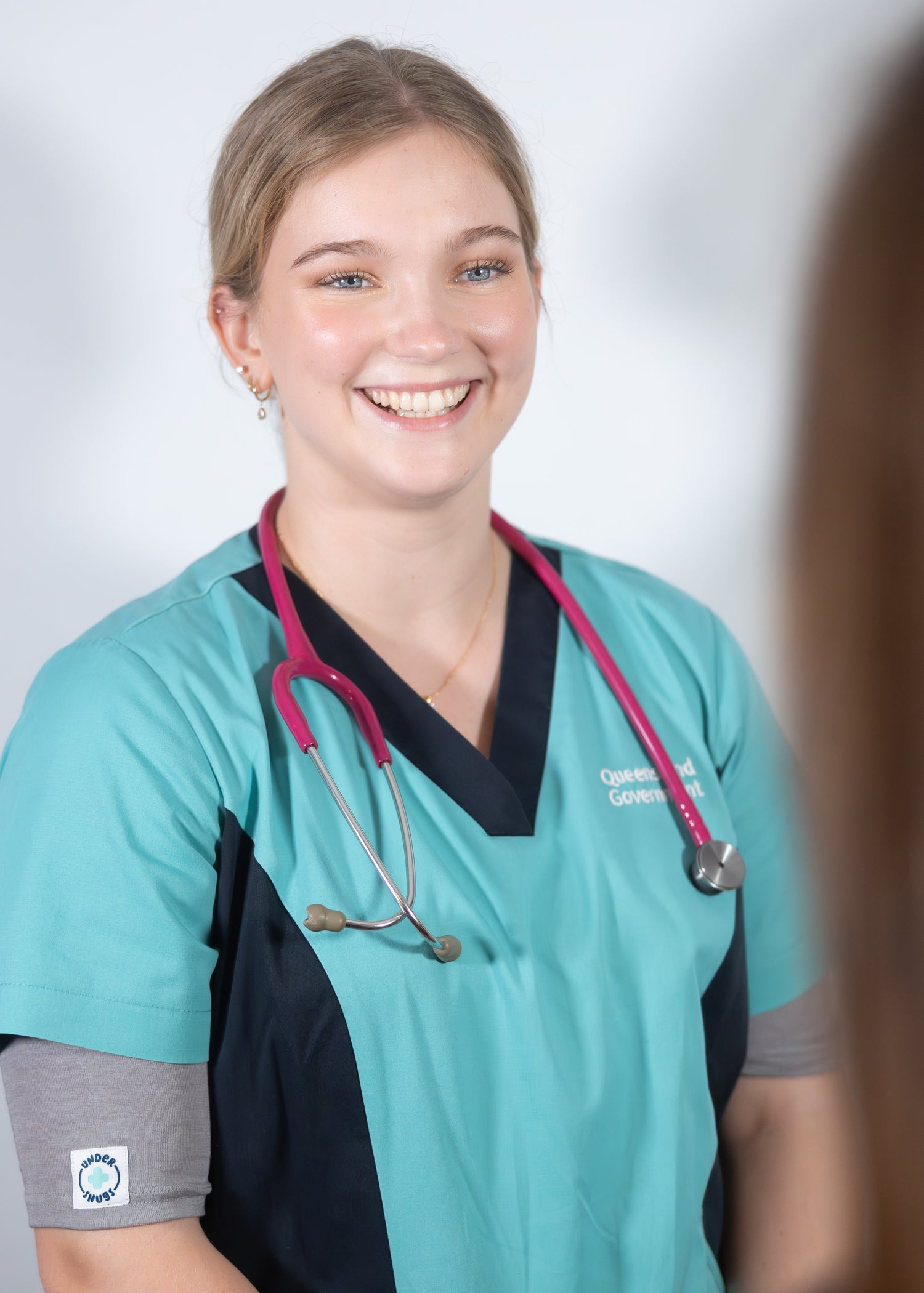 Young nurse smiling & wearing grey marle Under Snugs under a bright teal hospital top with a pink stethoscope around her neck