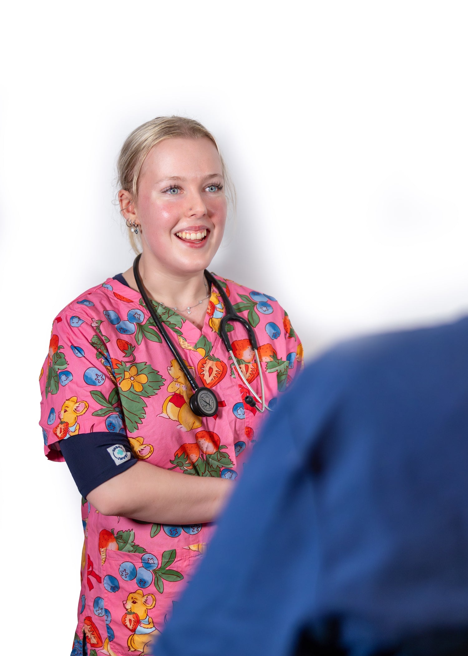 Young nurse smiling whilst talking with her patient, wearing navy blue Under Snugs under happy, pink kid-friendly patterned scrubs.