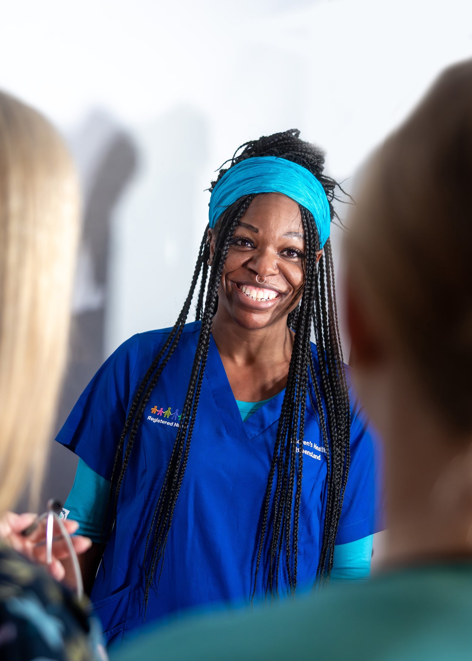 A young nurse smiling at her nurse colleagues as she chats with them, wearing peacock Under Snugs & bright blue scrubs